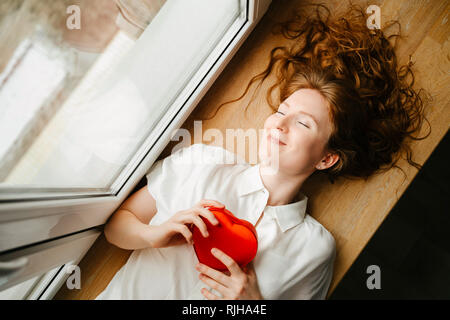 Belle jeune fille allongé par la fenêtre avec un cadeau pour la Saint-Valentin. Cheveux bouclés rouge. Banque D'Images