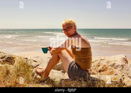 Femme, assis sur la roche par la mer et boire une tasse de café, le camping, Portbail, Normandie, France Banque D'Images