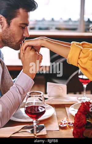 Proposition romantique. Jeune homme embrasse les mains de sa petite amie au restaurant. Des roses rouges et des bougies sont posé sur la table près du vin rouge. Portrait pho Banque D'Images