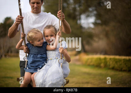 Deux petites filles souriant s'amuser soit poussé sur un arbre swing par leur père. Banque D'Images