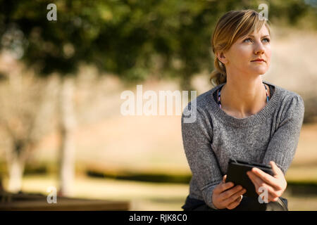 Jeune femme assise dans un jardin ensoleillé a l'air de sa tablette numérique et regarde pensivement dans la distance. Banque D'Images