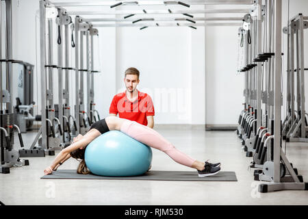 Jeune femme qui s'étend sur une boule de forme physique avec l'homme formateur pendant le traitement de la colonne vertébrale à la réhabilitation de sport Banque D'Images
