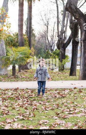 Enfant habillé en vêtements chauds à jouer avec l'automne les feuilles tombées dans un parc. Banque D'Images