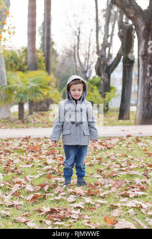 Enfant habillé en vêtements chauds à jouer avec l'automne les feuilles tombées dans un parc. Banque D'Images