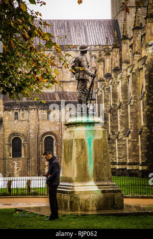 Winchester, Hampshire. Homme debout par un mémorial de guerre à la recherche à un téléphone mobile avec des arbres et de la cathédrale de Winchester aussi en vue. Banque D'Images