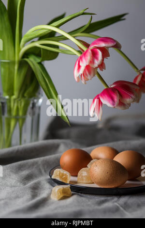 Les oeufs bouillis brown sur une assiette, à côté les tulipes dans une tasse en verre transparent sur fond gris dans une lumière et dans soft focus Banque D'Images