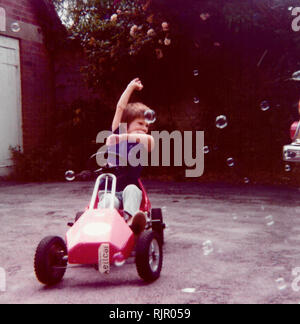 1970 - Un jeune garçon dans sa voiture à pédale zooms par bulles dans son jardin. Angleterre vers 1975. Banque D'Images