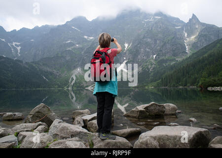 Une femme se tient sur la rive rocheuse du lac Morskie Oko (oeil de la mer) en prenant une photo avec son téléphone portable dans le Parc National des Tatras, Pologne Petite Vovo Banque D'Images
