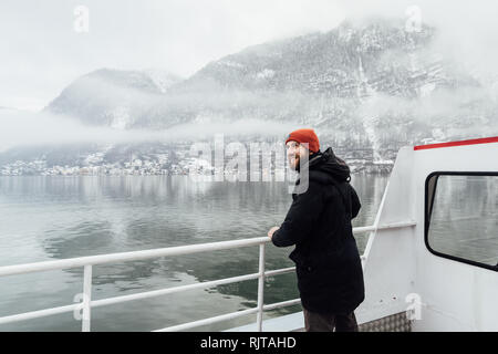 Homme barbu à chapeau orange debout sur un pont transbordeur sur le chemin de l'Hallstatt, Autriche. Vue d'hiver sur le lac et montagnes des Alpes. Banque D'Images