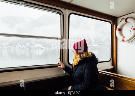 Woman in red hat assis à l'intérieur le traversier sur la façon d'Hallstatt, Autriche. Vue d'hiver sur le lac et montagnes des Alpes. Banque D'Images