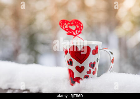 L'amour au temps de Noël, café concept, mug et coeur rouge décoration sur un balcon de la neige.Cup avec cœur et une boisson chaude enveloppée dans un foulard dans la neige Banque D'Images