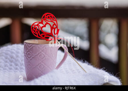 L'amour au temps de Noël, café concept, mug et coeur rouge décoration sur un balcon de la neige.Cup avec cœur et une boisson chaude enveloppée dans un foulard dans la neige Banque D'Images