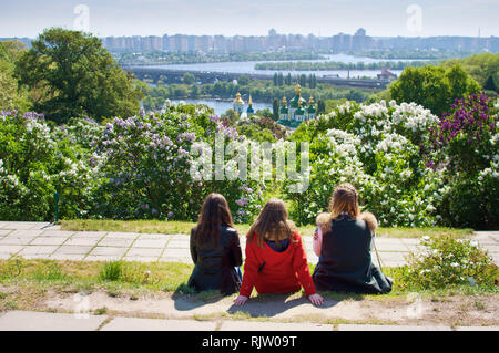 Trois jeunes filles assises sur le sol avec le dos tourné et à la recherche à dômes du monastère Vydubychi parmi les buissons de lilas en fleurs et feuillage vert ne Banque D'Images