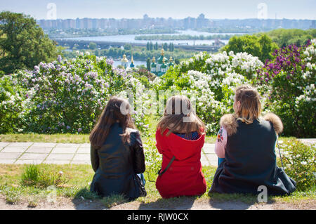 Trois jeunes filles assises sur le sol avec le dos tourné et à la recherche à dômes du monastère Vydubychi parmi les buissons de lilas en fleurs et feuillage vert ne Banque D'Images