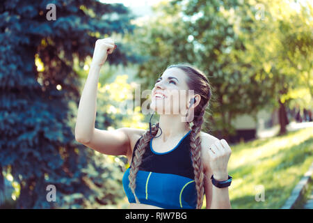 Fille sportive acclamations, gagnant. Femme chez sports cheerful avec bras tendus poings pompage smiling crier de joie à l'extérieur, dans un parc verdoyant sur backgroun Banque D'Images