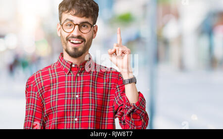 Beau jeune homme portant des lunettes au fond isolé montre de l'index jusqu'à une bonne idée. Quitté et heureux. Numéro un. Banque D'Images