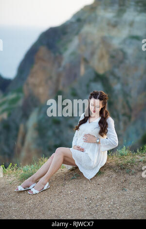 Femme enceinte posant assis sur la montagne falaise portant robe de pentecôte aérée avec bleu de la mer sur l'arrière-plan. Banque D'Images