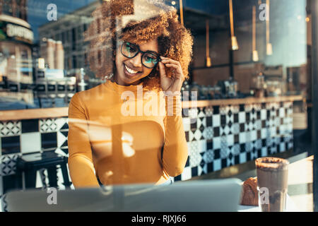 Smiling afro american woman working on laptop assis dans un café. Cheerful woman in eyeglasses sitting in a restaurant looking at a laptop vu Banque D'Images