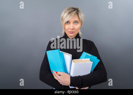 Portrait d'une blonde de la quarantaine, femme d'affaires, enseignant, entraîneur dans un chemisier noir et des jeans et lunettes turquoise avec des livres dans leurs mains sur un background gris Banque D'Images