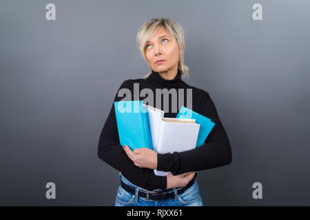 Portrait d'une blonde de la quarantaine, femme d'affaires, enseignant, entraîneur dans un chemisier noir et des jeans et lunettes turquoise avec des livres dans leurs mains sur un background gris Banque D'Images