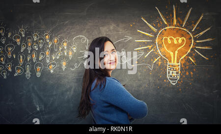 Jeune femme ingénieuse pensée positive des élèves des idées de rassemblement dans un grand tableau noir sur l'ampoule du feu arrière-plan. La créativité Genius concept de l'éducation. Banque D'Images