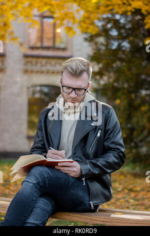 Smart Guy écrivain dans les tenues prend des notes sur un ordinateur portable et se repose sur un banc de parc Banque D'Images