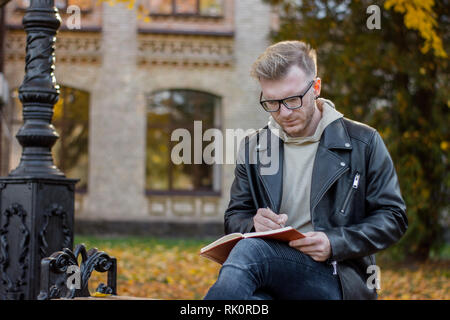 L'écrivain Guy attrayant dans les tenues prend des notes sur un ordinateur portable et se repose sur un banc de parc Banque D'Images