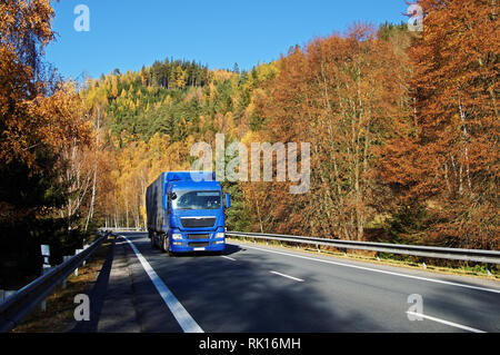 Camion bleu sur route asphaltée dans une vallée boisée au-dessous de la montagne, aux couleurs de l'automne flamboyant. Journée d'automne ensoleillée avec ciel bleu. Banque D'Images