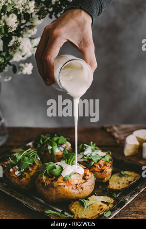 La main de l'homme de verser la crème sur les pommes de terre farcies au four avec du fromage, légumes et rucola sur table en bois. Fond rustique. Copy space Banque D'Images