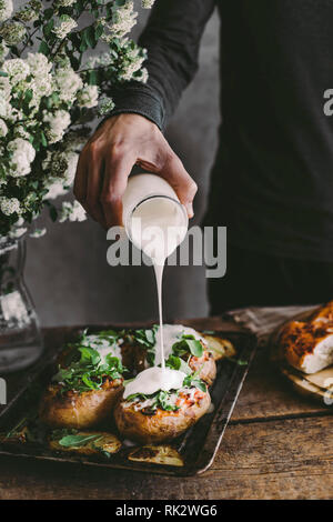 La main de l'homme de verser la crème sur les pommes de terre farcies au four avec du fromage, légumes et rucola sur table en bois. Fond rustique Banque D'Images