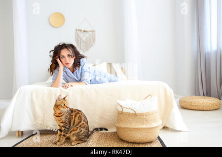 Portrait of attractive smiling woman in pyjama avec Bengal chat posant sur le lit chez lui Banque D'Images