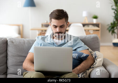 Serious man using laptop sitting on couch working at home Banque D'Images