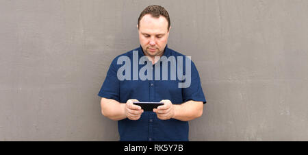 Le jeune homme de la chemise bleue utilise son téléphone intelligent en mur de béton copie espace. Banque D'Images