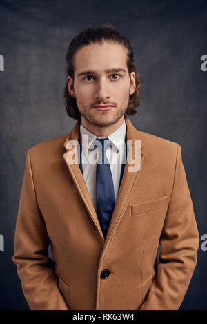 Portrait d'un jeune élégant bel homme aux longs cheveux brun clair non rasé en blazer et cravate bleu, looking at camera avec visage amical Banque D'Images