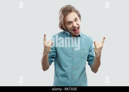 Portrait de screaming beau jeune homme blond aux cheveux longs en bleu chemise décontractée debout avec rock et signe à la caméra à crier. Piscine studio Banque D'Images