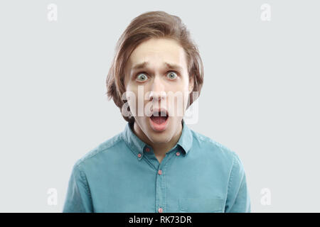 Portrait de choqué beau jeune homme blond aux cheveux longs en bleu shirt debout avec de grands yeux, bouche ouverte, surpris face à la caméra à l'intérieur. s Banque D'Images