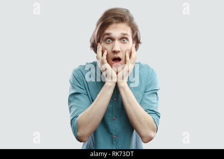 Portrait de surpris beau jeune homme blond aux cheveux longs en bleu shirt debout avec de grands yeux holding hand sur le visage et à visage choqué avec indo. Banque D'Images