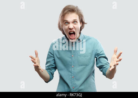 Portrait de crier en colère beau jeune homme blond aux cheveux longs en bleu chemise décontractée debout avec les bras levés, agressif looking at camera . indoo Banque D'Images