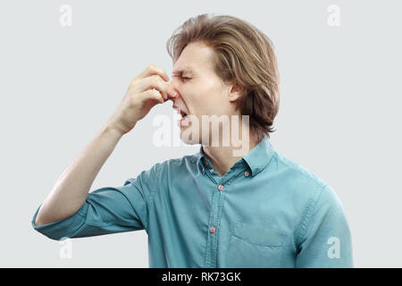 Portrait de profil Vue de côté malheureux beau jeune homme blond aux cheveux longs en bleu chemise décontractée debout pinçant le nez parce qu'une mauvaise odeur. Banque D'Images