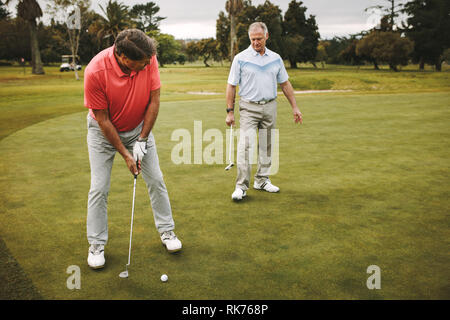 Golfeur aîné sur putting green sur le point de prendre la photo. Homme joueur de golf putting on vert avec deuxième joueur debout à l'arrière. Banque D'Images