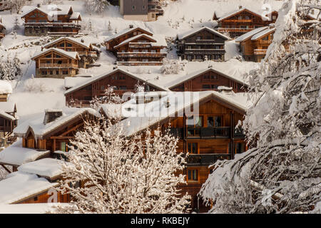 Derrière les arbres, vue sur un groupe de lumière recouvert de neige ski chalets en bois brun foncé dans une station de ski dans les alpes françaises. Banque D'Images