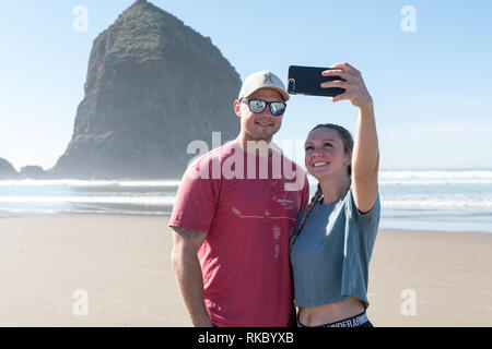Un jeune couple de touristes prennent vos autoportraits en face de l'Haystack Rock à Cannon Beach, Oregon, USA. Banque D'Images