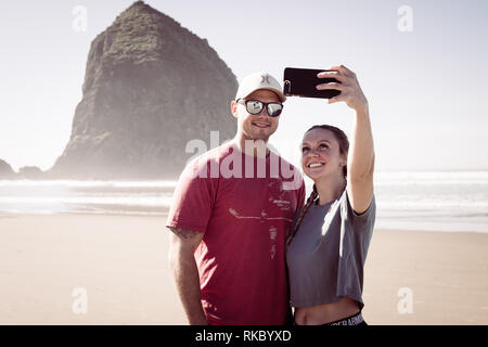 Un jeune couple de touristes prennent vos autoportraits en face de l'Haystack Rock à Cannon Beach, Oregon, USA. Banque D'Images