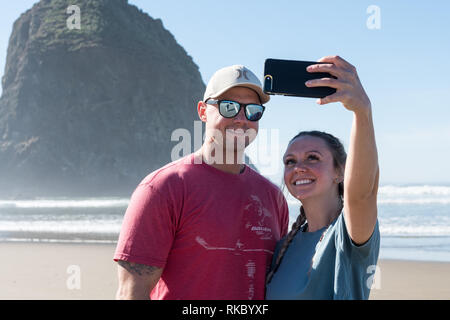 Un jeune couple de touristes prennent vos autoportraits en face de l'Haystack Rock à Cannon Beach, Oregon, USA. Banque D'Images