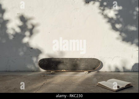 Skateboard appuyée sur mur blanc à côté d'un livre avec un escargot sur le dessus. Soirée ensoleillée en lieu naturel avec l'ombre des arbres dans le mur. Banque D'Images