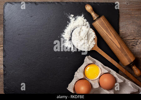 Ingrédients et ustensiles pour la cuisson. Avec une cuillère de farine, d'œufs, de rouleau à pâtisserie sur un arrière-plan en ardoise noire. Vue de dessus, selective focus, copy space Banque D'Images