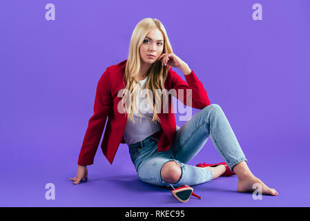 Jeune femme intéressée à Red Jacket assis sur le plancher sur fond violet Banque D'Images