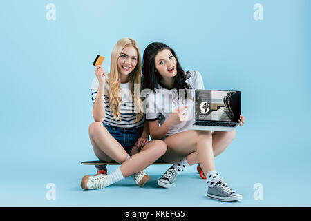 Smiling girls holding laptop avec booking site sur écran et une carte de crédit tout en restant assis sur longboard sur fond bleu Banque D'Images