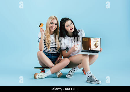 Les filles sur longboard holding laptop avec des billets en ligne site sur écran et de la carte de crédit sur le fond bleu Banque D'Images