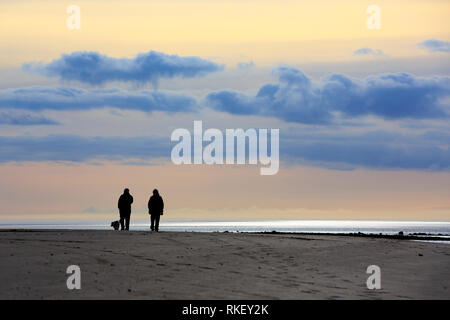 Ferryside, Carmarthenshire, Pays de Galles, Royaume-Uni. Lundi 11 février 2019. Un couple prendre leur chien pour une promenade sur la plage de Ferryside comme le soleil commence à définir sur un ressort-comme le jour dans l'ouest du pays de Galles. Credit : Gruffydd Thomas/Alamy Live News Banque D'Images